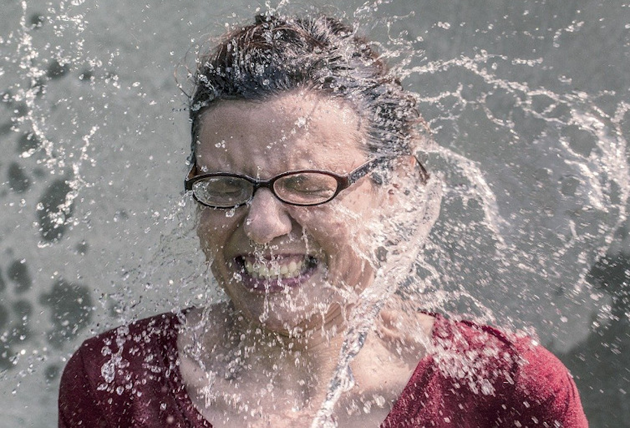 Das Bild zeigt eine Frau mit Brille, der ein Schwall Wasser ins Gesicht fällt.