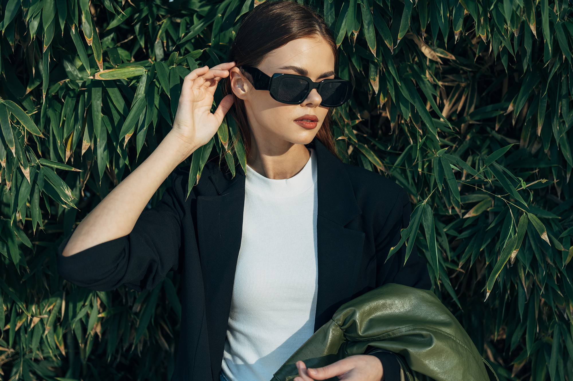Das Bild zeigt eine Frau mit Sonnenbrille vor einer grünen Hecke.